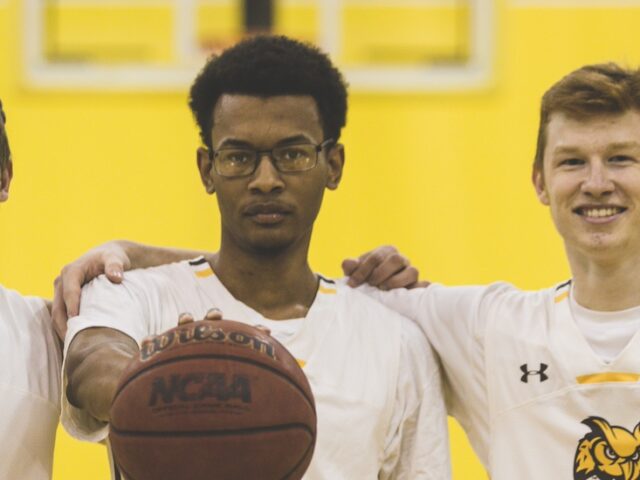 Three men's basketball players stand shoulder to shoulder looking at the camera, with the player in the middle palming a basketball and holding it out in front of him.