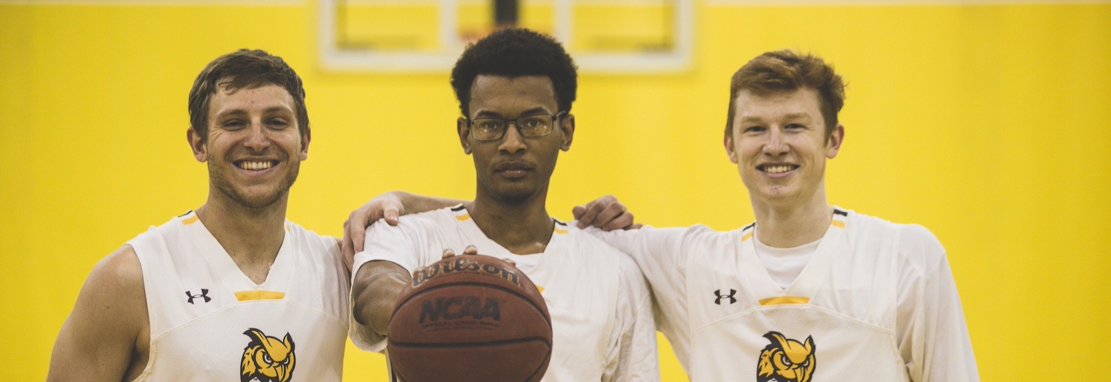 Three men's basketball players stand shoulder to shoulder looking at the camera, with the player in the middle palming a basketball and holding it out in front of him.