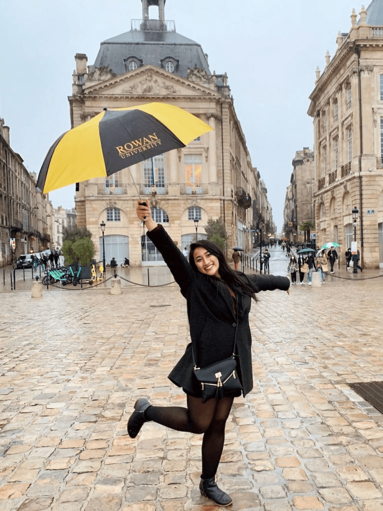 Rowan University alumna Stephanie Ibe holds a Rowan umbrella high above her head, with her right leg kicking backward in a casual way, in Vieille Ville (Bordeaux).
