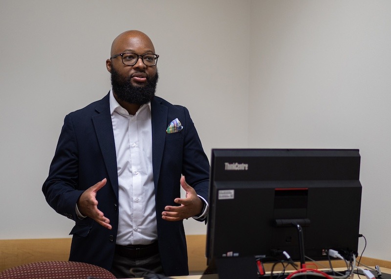 A Rowan University graduate teaches in front of a classroom, using his hands to speak and with a computer monitor on the desk in front of him. 