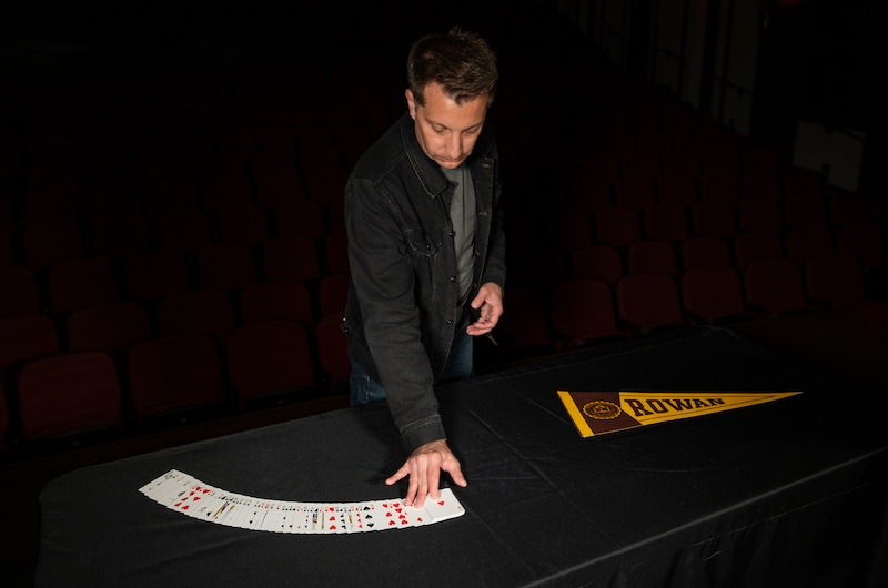 Magician and Rowan marketing alumnus David Corsaro looks down at a table where he has spread a deck of cards, next to a Rowan banner. 