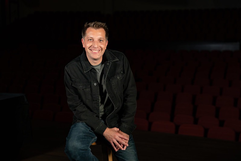 Magician and Rowan marketing alumnus David Corsaro smiles for a portrait, while sitting casually on a stool, with his hands loosely clasped. 