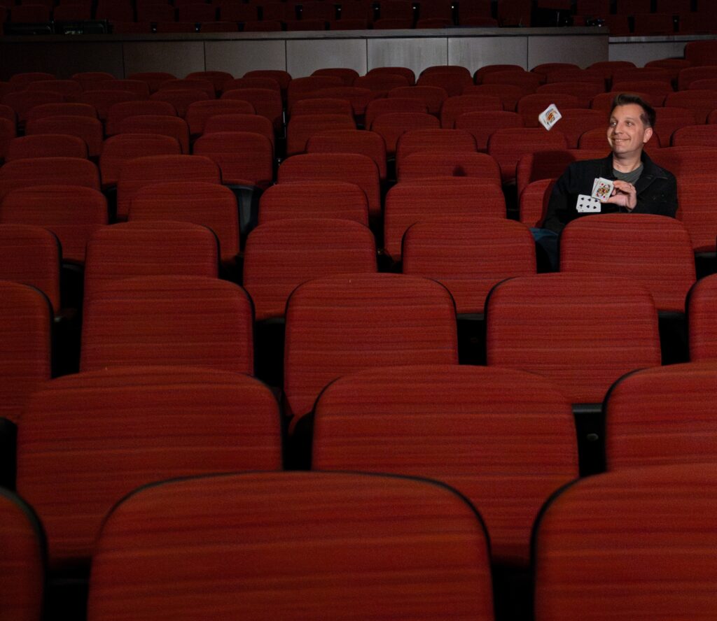 Magician and Rowan marketing alumnus David Corsaro playfully throws cards in the air while sitting alone in empty theatre seats. 