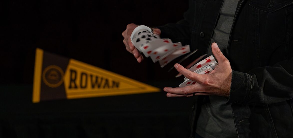 A close up of the hands of magician & Rowan marketing alumnus David Corsaro as he shuffles a deck of cards while next to a Rowan banner.