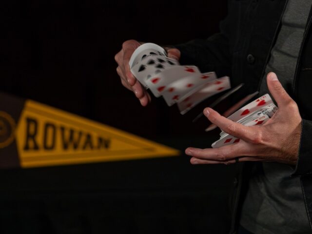 A close up of the hands of magician & Rowan marketing alumnus David Corsaro as he shuffles a deck of cards while next to a Rowan banner.