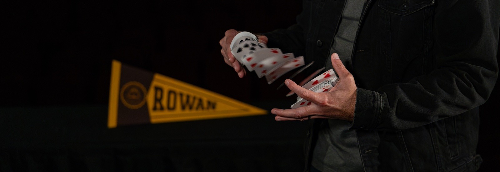 A close up of the hands of magician & Rowan marketing alumnus David Corsaro as he shuffles a deck of cards while next to a Rowan banner.