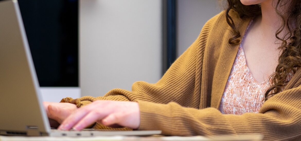 A close up of hands working on a laptop as the student wears a light brown cardigan.
