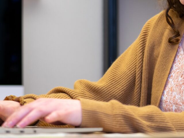 A close up of hands working on a laptop as the student wears a light brown cardigan.