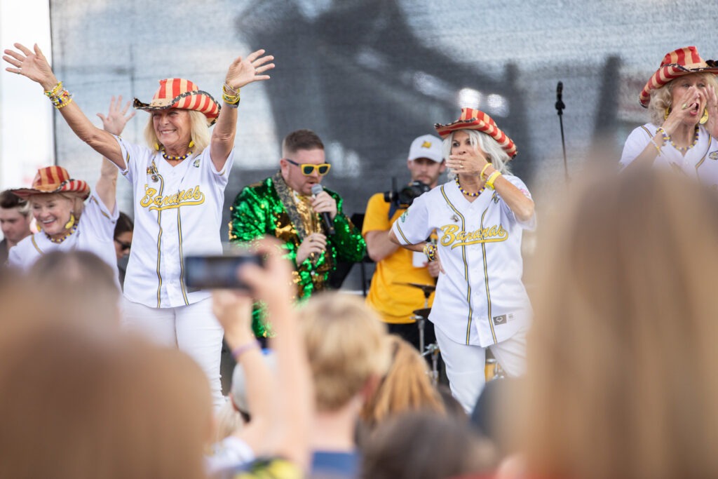Nana Karen is seen arms outstretched with a smile on her face during a performance, with fellow dancers and fellow dancers around her and the crowd filming them on their phones. 