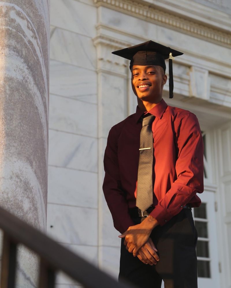 Devón Coles stands with his hands clasped in front of him, wearing a dress shirt and tie and his college graduation cap and tassel. 