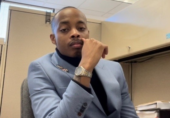 Devón Coles sits at his desk, wearing a gray suit jacket, his hand raised to his face. 