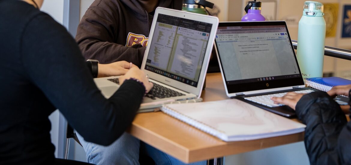 A close up of the hands and laptops of three online students working side by side.