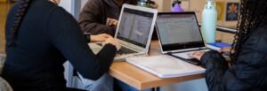 A close up of the hands and laptops of three online students working side by side.