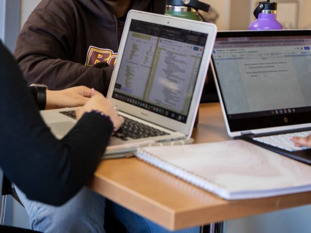 A close up of the hands and laptops of three online students working side by side.