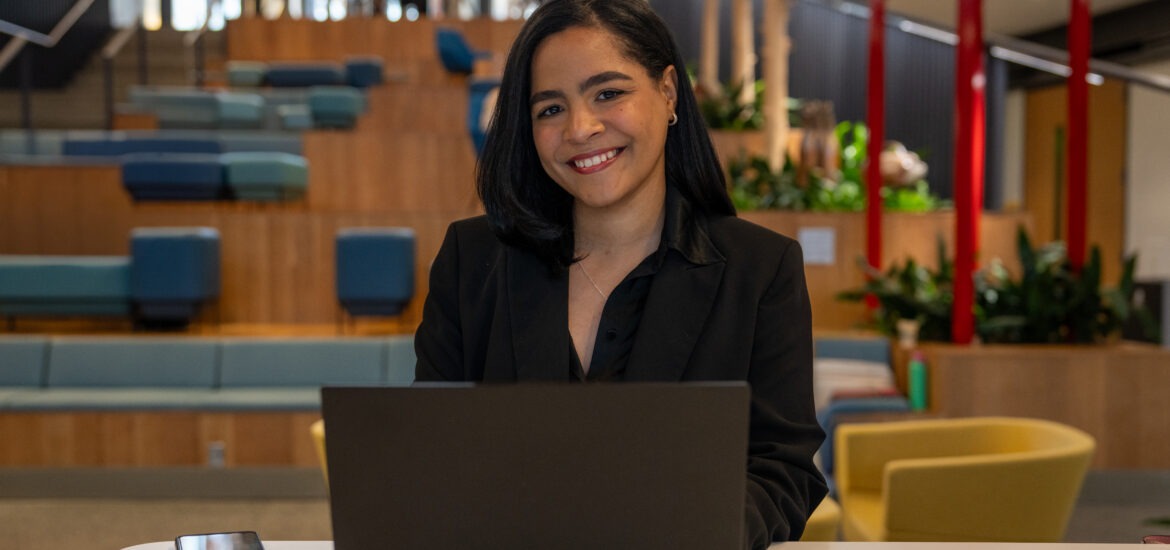 Flerida V., online BA in Liberal Studies student, sitting down at table smiling with laptop open.