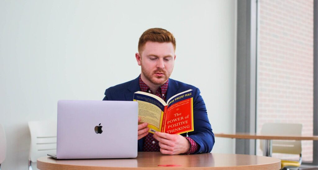 Online male psychology student sitting at table with laptop open reading textbook.