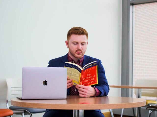 Online male psychology student sitting at table with laptop open reading textbook.