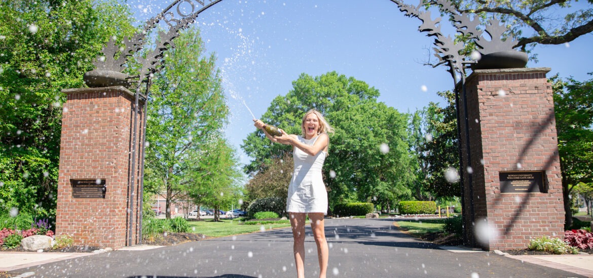 A Rowan University graduate in a white dress celebrates by popping champagne beneath the Rowan University arch on a sunny day, symbolizing achievement, joy, and new beginnings through Rowan’s online graduate programs.