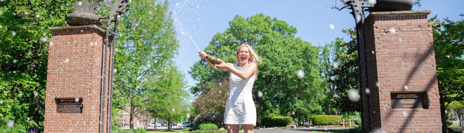 A Rowan University graduate in a white dress celebrates by popping champagne beneath the Rowan University arch on a sunny day, symbolizing achievement, joy, and new beginnings through Rowan’s online graduate programs.
