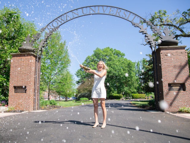 A Rowan University graduate in a white dress celebrates by popping champagne beneath the Rowan University arch on a sunny day, symbolizing achievement, joy, and new beginnings through Rowan’s online graduate programs.