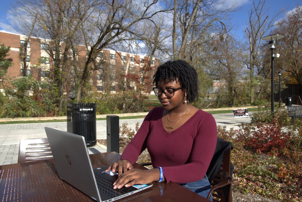Rowan chemistry major Layahna sits outdoors at a table outside the student center, working on her laptop, with a walkway, trees and a dorm behind her. 