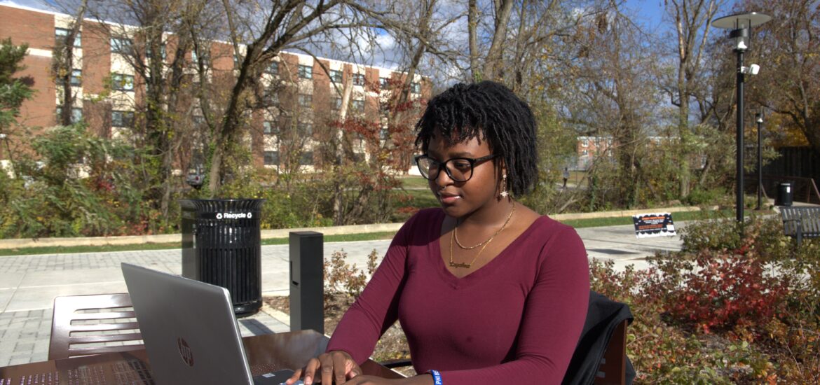 Student wearing glasses and a burgundy top works on a laptop at an outdoor table on Rowan University’s campus, with trees and campus buildings in the background.
