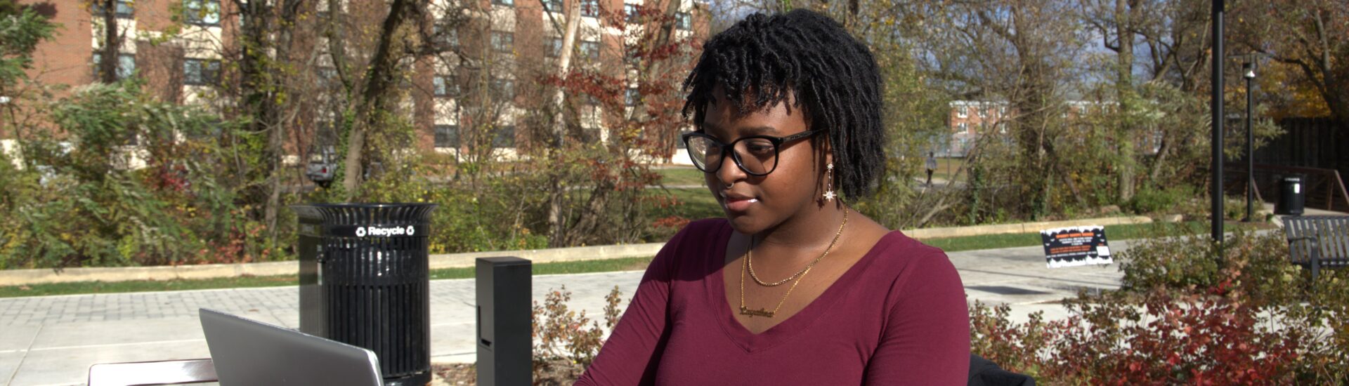 Student wearing glasses and a burgundy top works on a laptop at an outdoor table on Rowan University’s campus, with trees and campus buildings in the background.