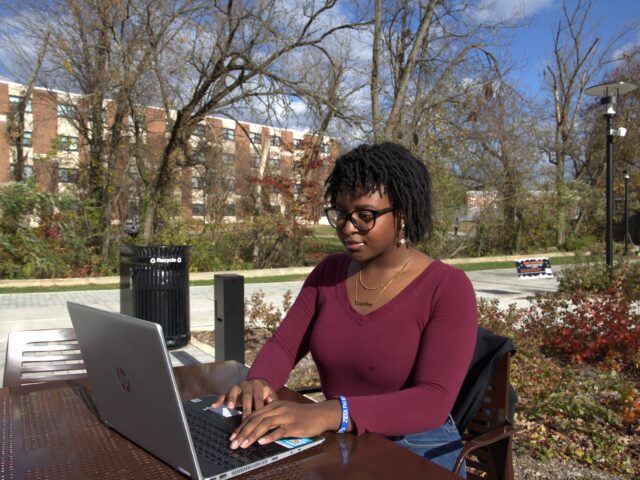Student wearing glasses and a burgundy top works on a laptop at an outdoor table on Rowan University’s campus, with trees and campus buildings in the background.