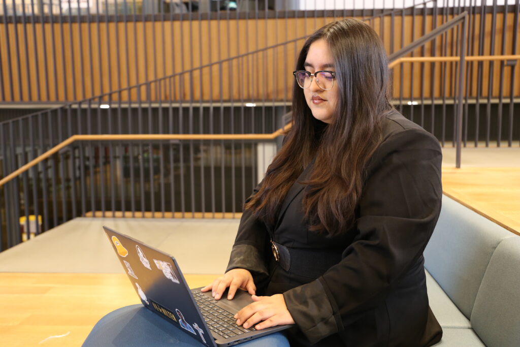 Student wearing glasses and a black blazer works on a laptop while seated at a table inside a modern Rowan University building.