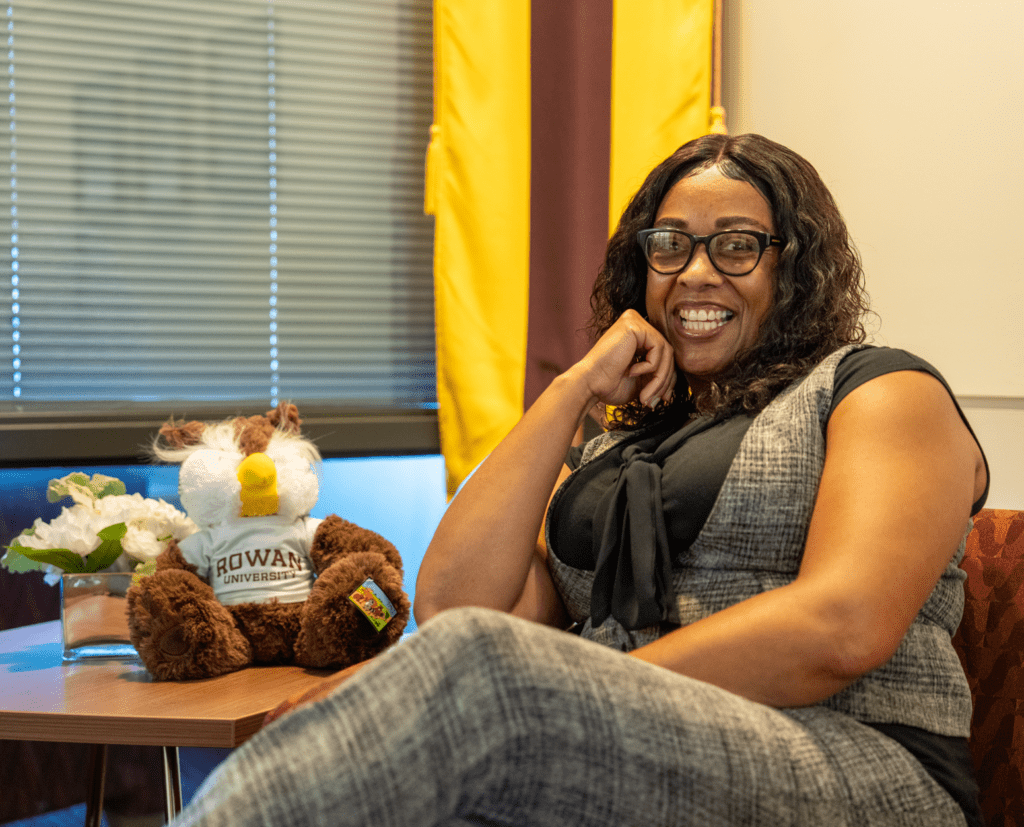 Rowan Online graduate smiling while sitting on couch with Rowan University stuffed animal in background.