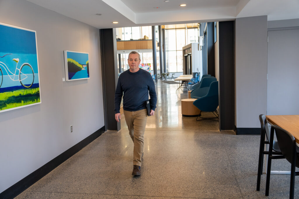 Bill Y., online bachelor's in respiratory therapy student walking down hallway with laptop in his hand, reflecting on his experience.