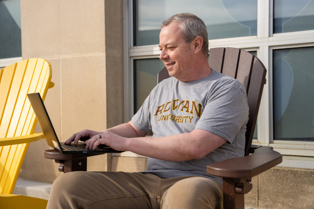 Bill Y., online bachelor's in respiratory therapist student sitting in brown chair outside completing online coursework on laptop with Rowan University t-shirt on.