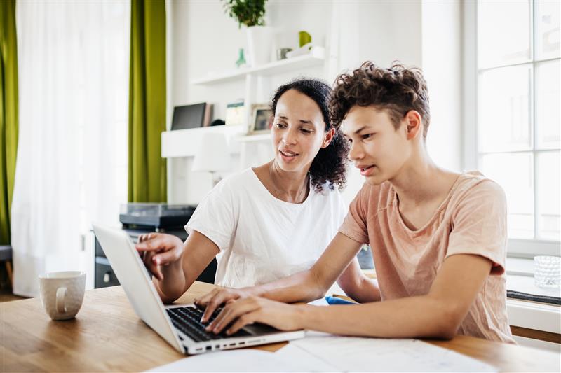 Mother sitting with high school senior son on laptop, providing guidance and support through the college decision-making process.
