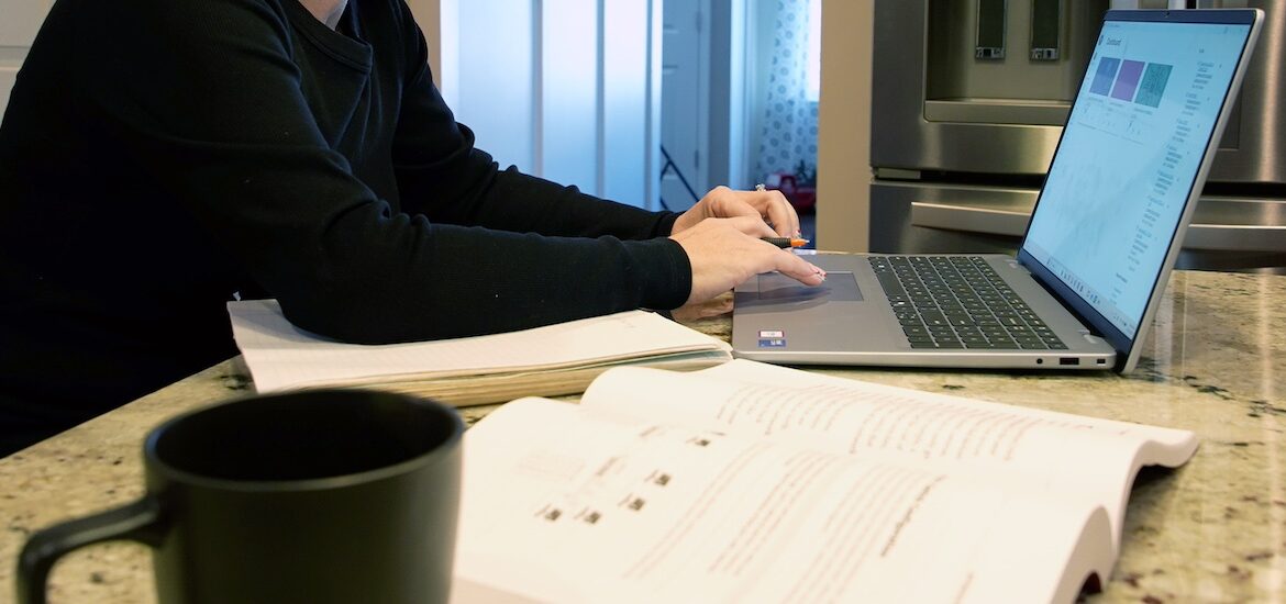 Rowan online master's in cybersecurity student Jennifer's workstation in her kitchen which includes her notebook, textbook and coffee mug, with Jennifer typing on her laptop.