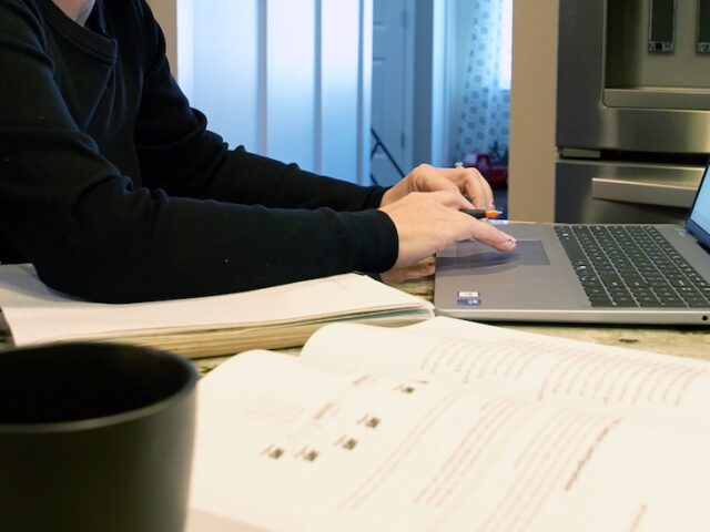 Rowan online master's in cybersecurity student Jennifer's workstation in her kitchen which includes her notebook, textbook and coffee mug, with Jennifer typing on her laptop.