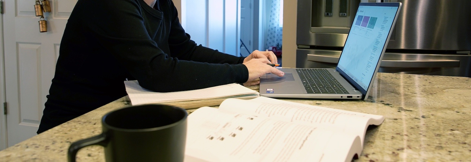 Rowan online master's in cybersecurity student Jennifer's workstation in her kitchen which includes her notebook, textbook and coffee mug, with Jennifer typing on her laptop.