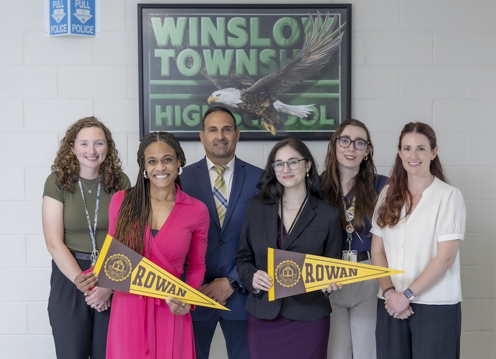 A group photo of the six educators, in front of a Winslow Township High School banner and holding Rowan penents in front of them.