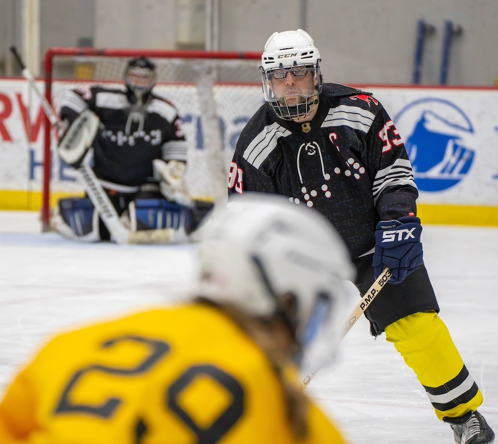 An action shot, Josh is seen over the head of his hockey opponent. He has a serious look on his face. The goalie behind him stands in the goalie box in a defensive position. 