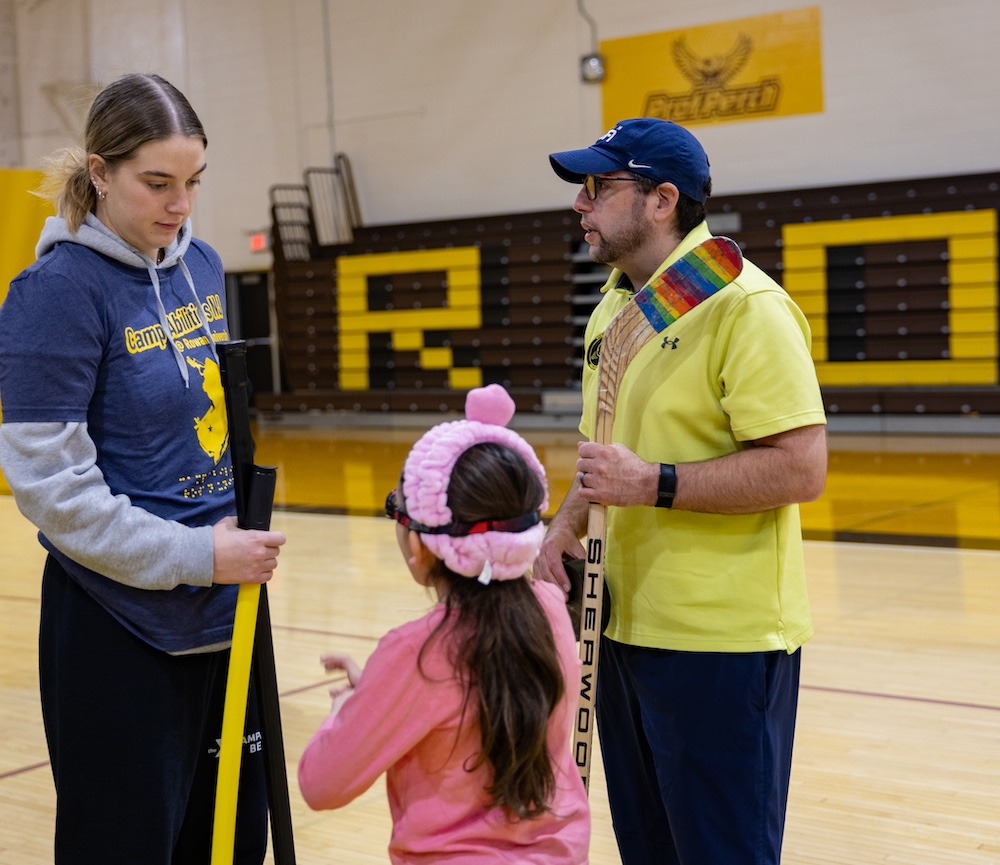 Master's of special education grad Josh Schneider stands in a Rowan University gymnasium, wearing a yellow collared shirt and holding a hockey stick. He is talking with a Camp Abilities leader and a little girl wearing all pink. 