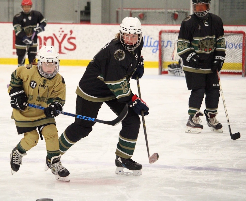 Three hockey players wearing dark uniforms play on the ice alongside a child who appears to be approximately 10 to 12 years old. 