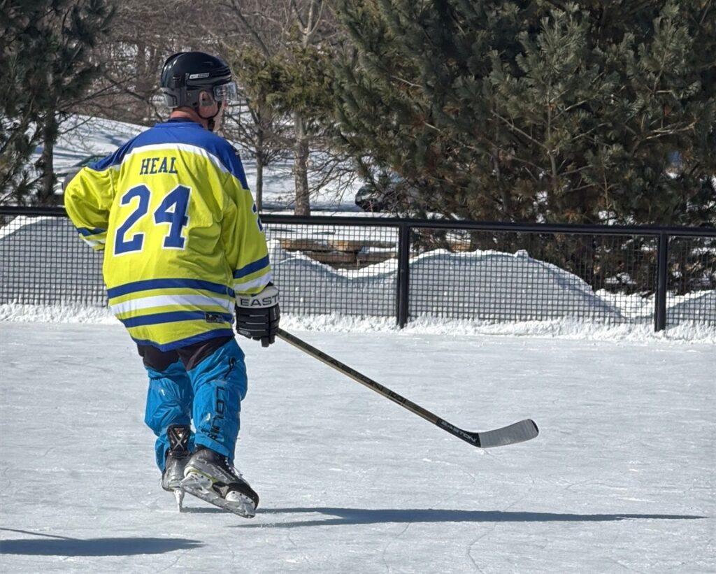 On an outdoor ice rink wth a fence around it and pine trees in the distance, a male hockey player skates while holding a hockey puck. 