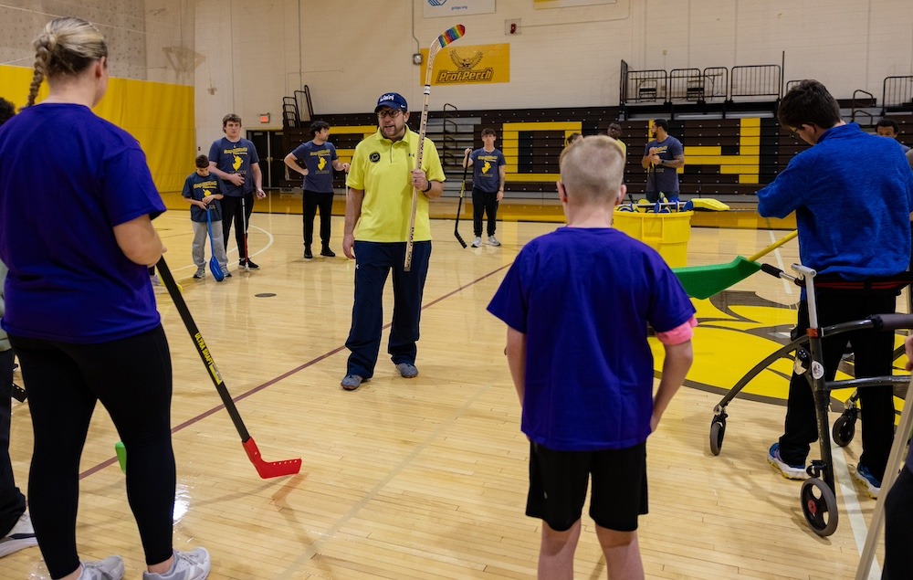 Josh Schneider stands in the center of a human-formed circle, in the Esby Gym at Rowan University. Around him are children and leaders from Campus Abilities, listening to Josh's instructions. 