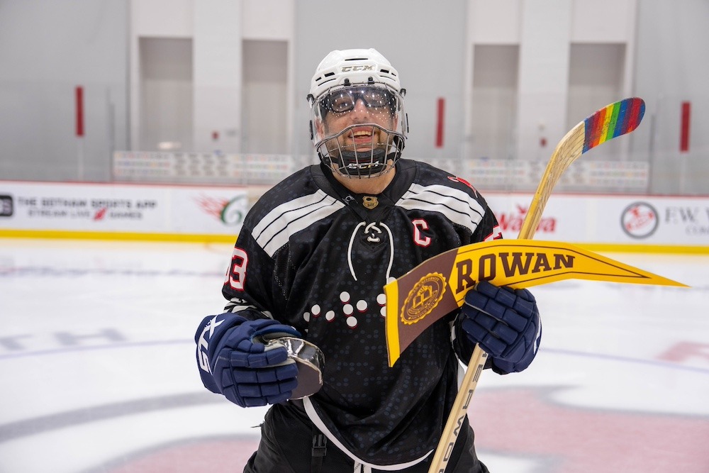 Josh Schneider poses for a portrait on the ice, wearing a black hockey uniform and a white helmet. He is holding a hockey puck in his right hand, and a hockey stick and a yellow and brown Rowan pennant in his left hand. 