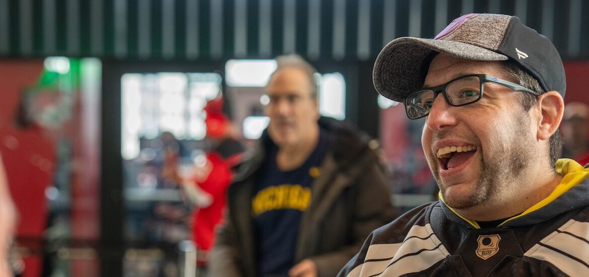 Rowan master's of special education alumnus Josh Schneider turns slightly to the right, smiling as he is speaking to someone. He is wearing a hockey uniform, glasses and a baseball cap.