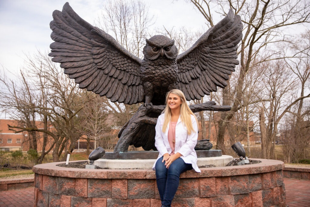 Pharmaceutical sciences master's student Holli-Joi sits in front of the owl statue on Rowan's campus. 