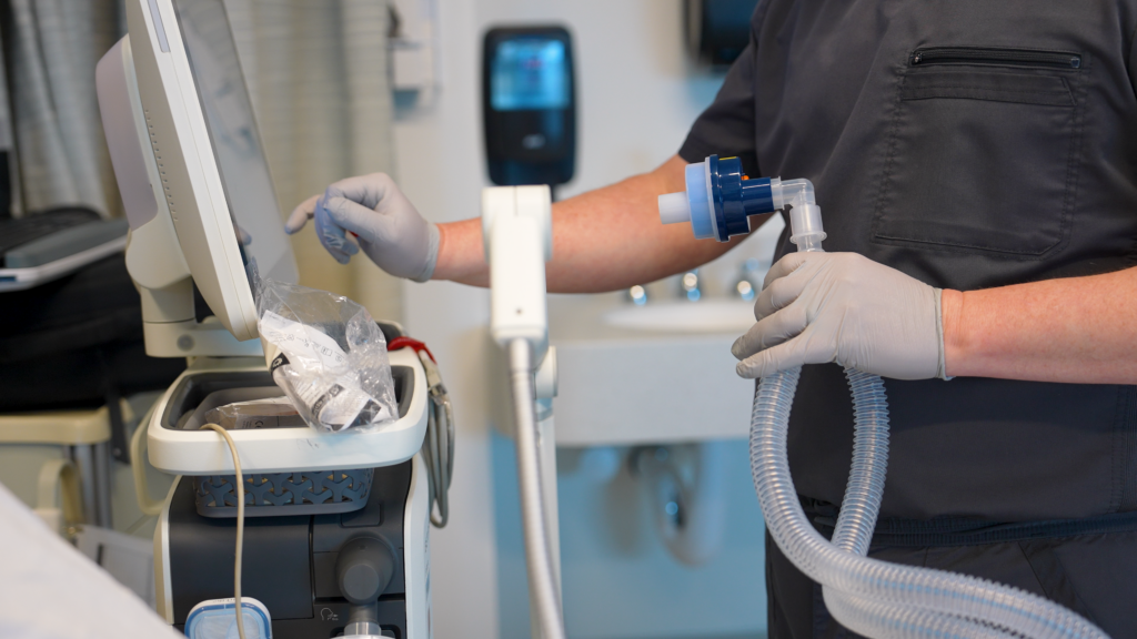 Respiratory therapist using clinical equipment to support patient breathing in a healthcare setting.