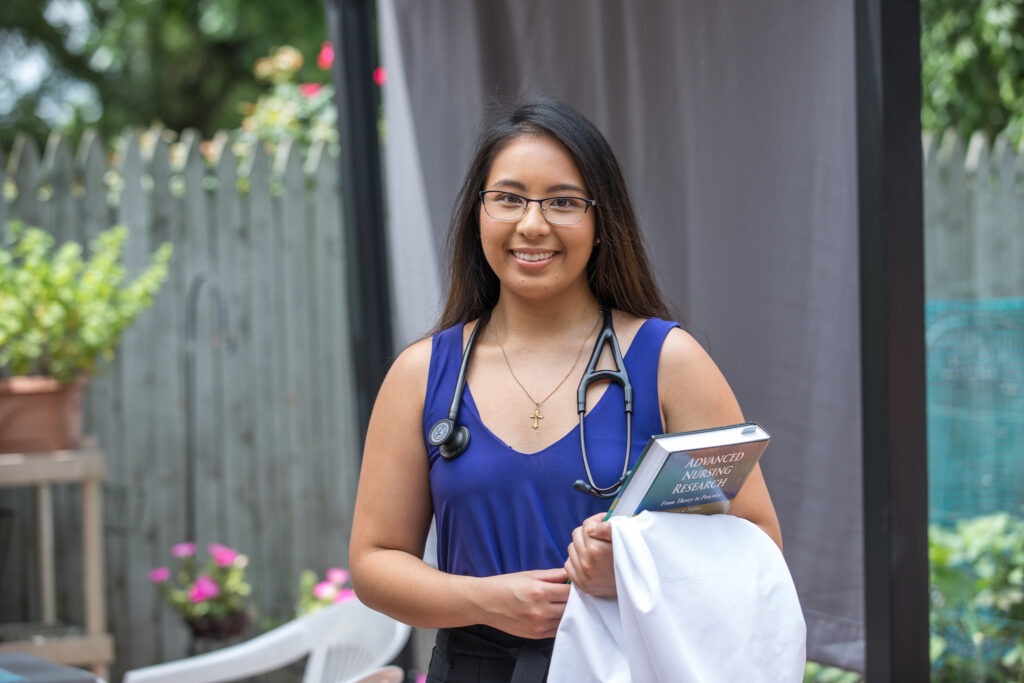 A Rowan master of science student stands in her garden, ready for school with a white lab coat draped over her forearm, a college textbook in hand, and a stethoscope around her neck. 