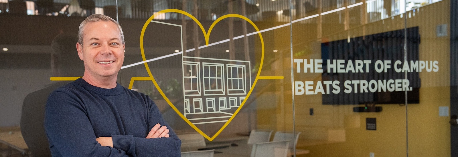 Standing in front of glass etched with the words "the heart of campus" a Rowan student stands smiling, his arms crossed in front of him.