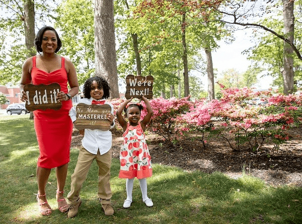 A Rowan master's graduate holds a sign that says "I did it!" as her young son next to her holds a sign that says "No, mom, you mastered it!" and her toddler daughter next to him, holding a sign above her head that says, "We're next!"
