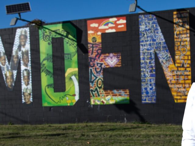 A Rowan student stands with her hand on her hip, looking right toward the sun with a mural behind her that paints the word Camden.
